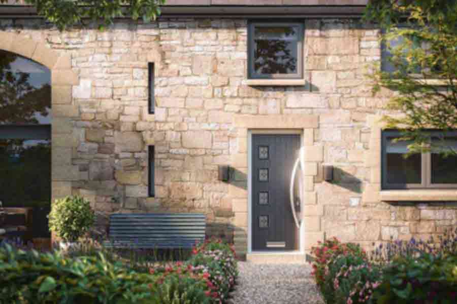 Contemporary stone house entrance featuring a dark composite front door with square glass inserts and curved handle, flanked by wall lights and surrounded by landscaped flower beds and gravel path.