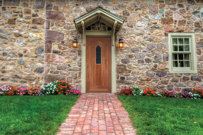 Stone house with wooden door and window, surrounded by flowers and a brick path.